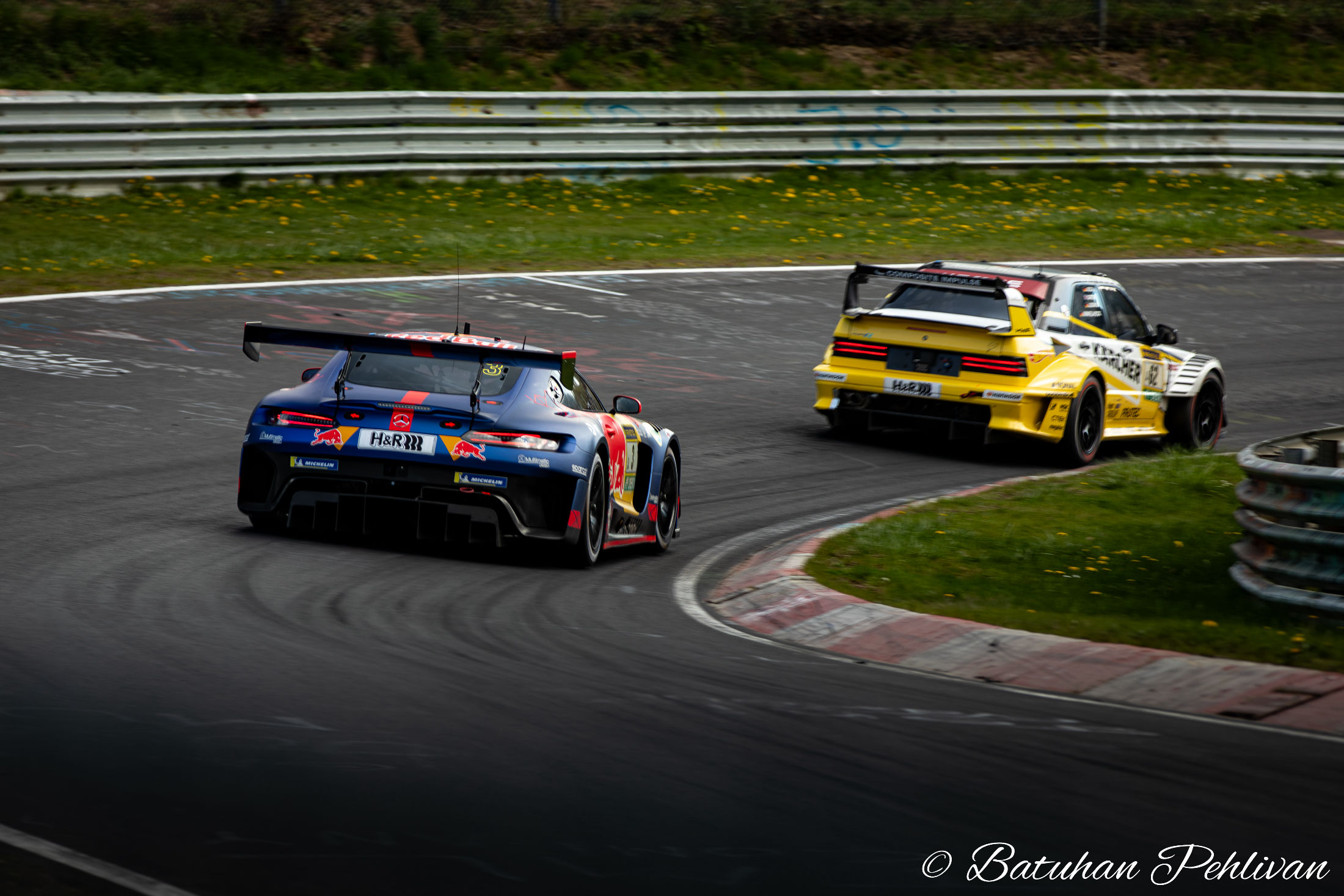 Two GT race cars battling through a corner on a racing circuit, showing close motorsport action and trackside atmosphere.